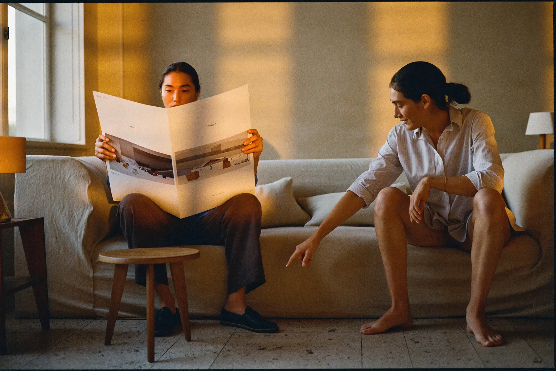 Two designers on a linen sofa in a sunlit studio, one holding large printed proofs, the other pointing at a detail.